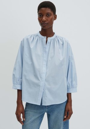 Vrouw draagt een losse, lichtblauwe blouse met driekwart mouwen, knoopsluiting en een ronde kraag, gecombineerd met een blauwe spijkerbroek, staand tegen een effen achtergrond.