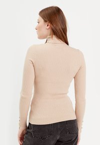 Jersey acanalado de color beige claro con cuello alto, mangas largas y pequeños botones decorativos en los puños. Tejido de punto texturizado con silueta ajustada.
