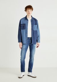 Camicia in denim con tonalità di blu miste, maniche lunghe, chiusura con bottoni e due tasche sul petto, abbinata a jeans blu e sneakers bianche.