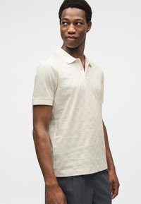 Ανοιχτό μπεζ κοντομάνικο πόλο με διακριτικό γεωμετρικό μοτίβο. Διαθέτει απαλό γιακά και πατιλέτα, φτιαγμένο από αναπνεύσιμο ύφασμα.