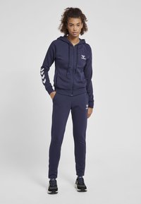 Mörkblå zip-up hoodie med dragsko, prydd med vitt logo och chevron-accenter på ärmarna, kombinerad med matchande åtsittande joggers.