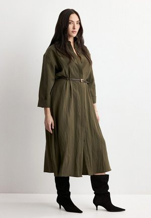 Femme debout dans une robe midi vert olive ceinturée à manches longues, portant des bottines à talons noires à bout pointu, sur un fond uni.