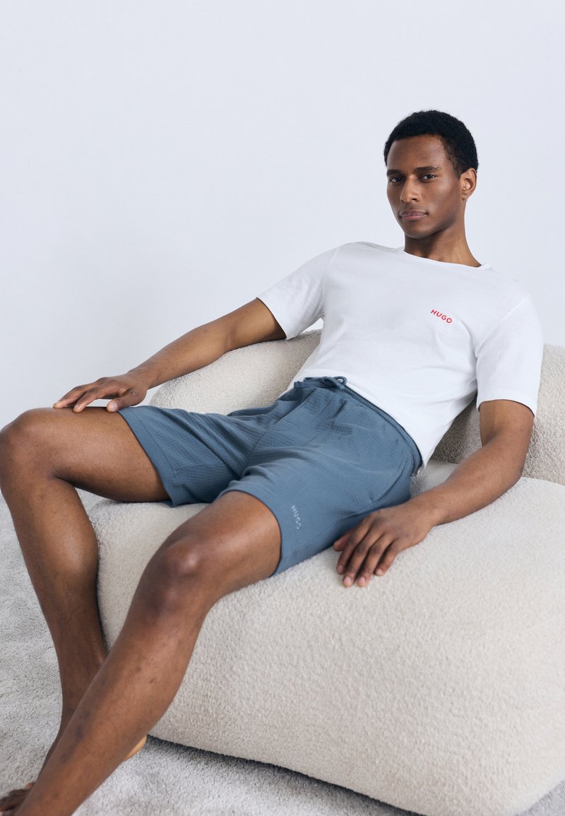 White short-sleeve shirt with red "HUGO" logo, paired with blue textured shorts. Model seated on a light-colored, plush surface.