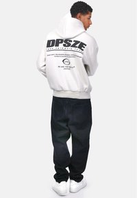 Un hoodie blanc avec un grand texte noir dans le dos, associé à un pantalon sombre et à des baskets blanches. Le hoodie possède un cordon de serrage et des poignets côtelés.