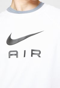 En person som bär en vit sweatshirt med grå krage, prydd med en svart Nike-swoosh-logotyp och ordet "AIR" tryckt på bröstet.