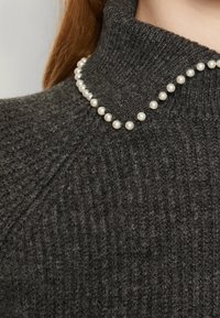 Maglione grigio scuro lavorato a maglia con collo alto e una fila di perle bianche lungo lo scollo, che mostra un tessuto strutturato.