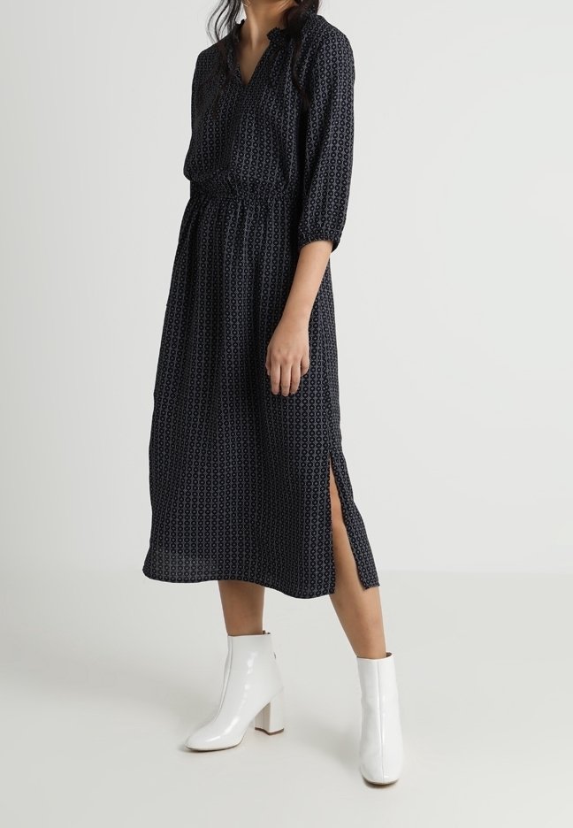 Femme portant une robe midi noire à motifs avec manches trois-quarts et fente latérale, associée à des bottines blanches.