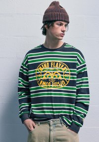 Prúhovaná dlhá mikina v tmavomodrej, zelenej a bielej farbe s žltým grafickým logom na hrudi. Má voľný strih a rebrovaný golier.