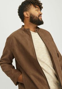 Chaqueta de ante marrón con cuello alto, cierre de cremallera en el frente y bolsillos laterales, combinada con una camisa ligera con textura por debajo.