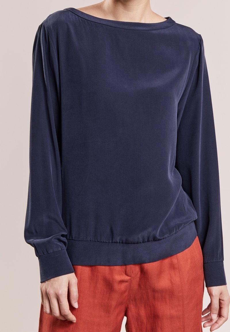 Personne portant un chemisier bleu marine uni à manches longues avec de larges poignets et un pantalon couleur rouille sur un fond neutre.