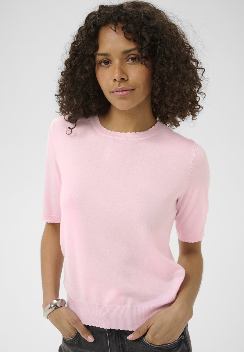 Vrouw met krullend haar, gekleed in een lichtroze gebreide top met korte mouwen, staand met handen in de zakken van een zwarte broek tegen een effen achtergrond.