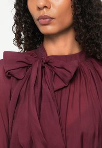 Femme aux cheveux bouclés portant un chemisier bordeaux avec un grand nœud noué au niveau du col et des détails en tissu froncé.