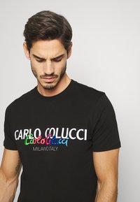 Sort bomulds t-shirt med farverig broderet tekst "CARLO COLUCCI" og "MILANO ITALY." Korte ærmer, rund hals, tætsiddende design.