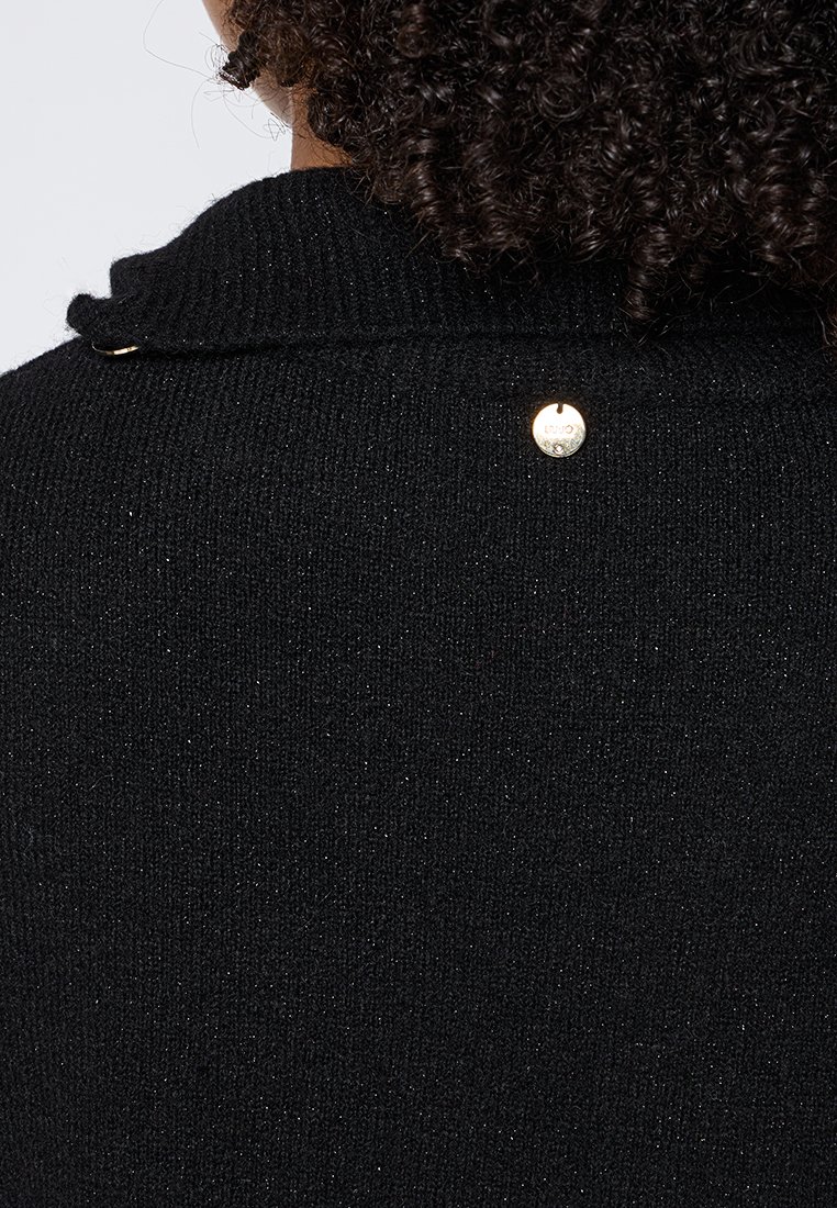 Capelli ricci raccolti, indossa un maglione nero tessuto con una piccola emblema d'oro rotonda vicino al colletto posteriore.