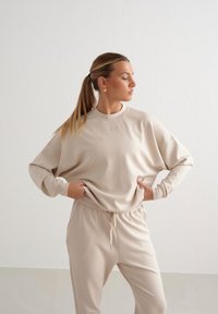 Felpa beige in pile con scollo a girocollo e spalle abbassate, abbinata a pantaloni della tuta tapered coordinati con vita elastica.