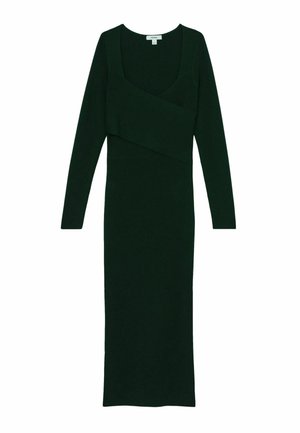 Mujer con cabello rizado y corto que lleva pendientes de oro y un vestido de manga larga verde oscuro con escote cuadrado sobre un fondo claro.