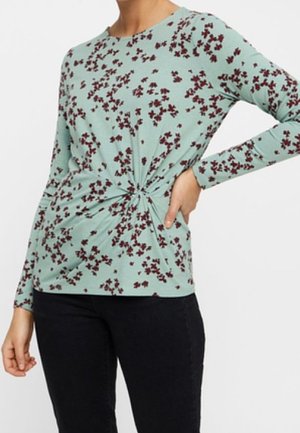 Femme portant un haut bleu clair à manches longues avec un petit imprimé floral rouge foncé et un détail torsadé à l'avant, associé à un pantalon noir.