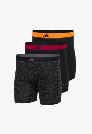 Oberkörperfreier Mann steht barfuß und trägt schwarze Boxershorts mit einem orangefarbenen Bund, auf dem ein schwarzes Adidas-Logo zu sehen ist.