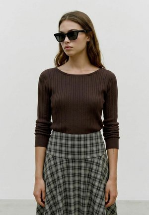 Femme portant des lunettes de soleil noires, un haut à manches longues en maille torsadée marron et une jupe taille haute à carreaux noirs et blancs, debout devant un fond uni.