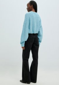 Hellblau strukturierter Strickpullover mit langen Ärmeln, kombiniert mit hochgeschnittenen schwarzen Schlagjeans. Schlichte schwarze Schuhe, sichtbare Gesäßtaschen.