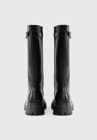 Botas de couro pretas até ao joelho com um acabamento texturizado, sola de borracha grossa, detalhe de fivela traseira e acabamentos em costura vertical.