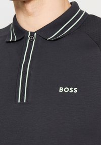 Polo-shirt sombre avec un col multirayé, une patte de boutonnage à deux boutons et des manches en mesh. Présente un logo "BOSS" de couleur vert clair sur la poitrine.