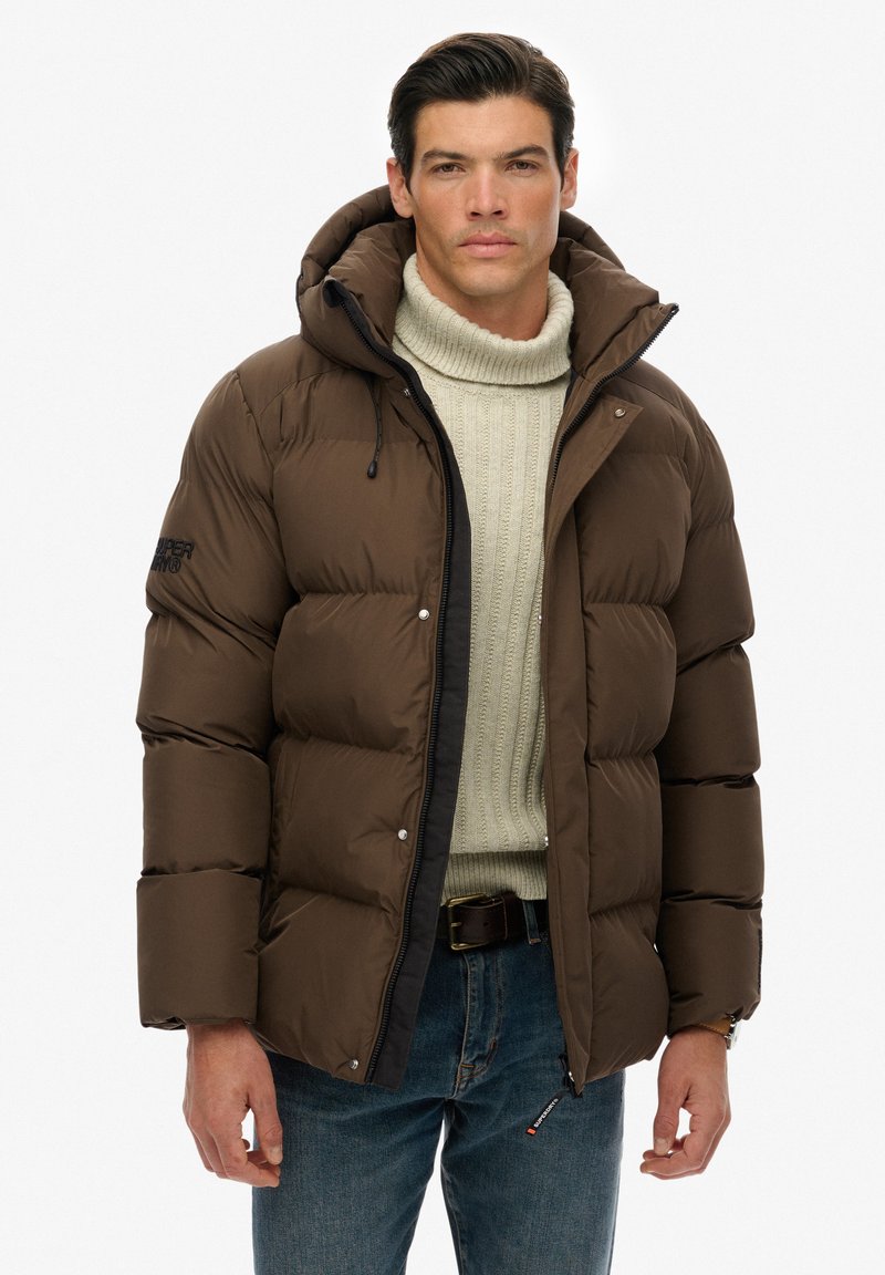 Chaqueta acolchada marrón con capucha, diseño acolchado, cremallera negra y logo en la manga izquierda. Lleva puesta sobre un suéter crema y jeans azules.