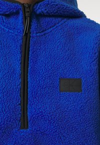 Pullover in pile blu con mezza zip, caratterizzato da una superficie texturizzata, zip nera e un'etichetta rettangolare nera.