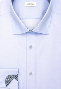 Camicetta azzurro chiaro con bottoni bianchi, colletto a punta e un tessuto a motivo blu contrastante all'interno dei polsini e del colletto.