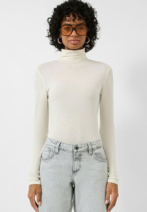 Jonge vrouw met krullend haar die grote bruine zonnebrillen draagt, een crèmekleurige coltrui met lange mouwen en lichtgrijze jeans met hoge taille.