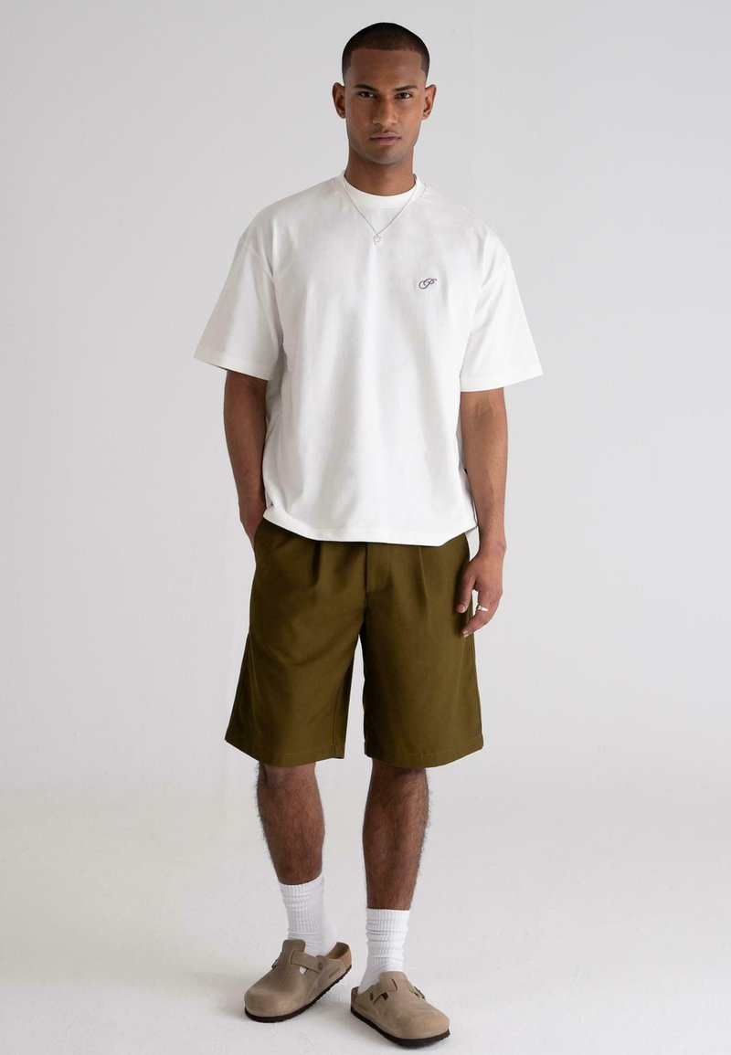 White cotton t-shirt with a small logo, paired with olive green shorts. Model wears beige clogs and white socks, standing on a plain background.
