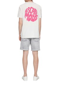 Weißes Baumwoll-T-Shirt mit einem großen, pinken "ESCAPE MOOD"-Grafik auf dem Rücken. Tragen mit grauen Jeansshorts, umgeschlagenen Säumen und weißen Turnschuhen.