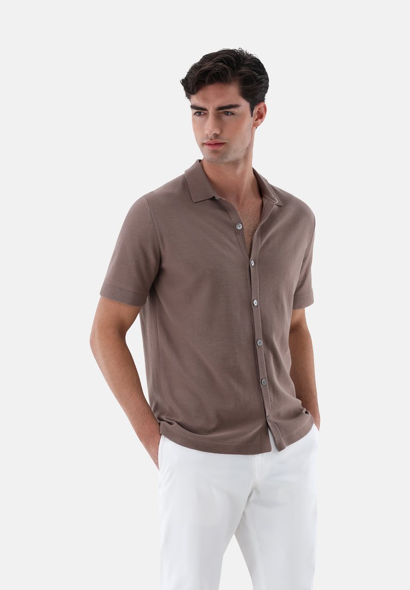 Camicetta a maniche corte marrone in maglia con colletto e bottoni frontali, abbinata a pantaloni bianchi. Tessuto morbido, vestibilità tailored.