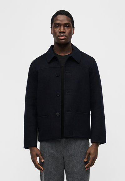 Άνδρας που φοράει μαύρο σακάκι με κουμπιά πάνω από μαύρο πουκάμισο και γκρι παντελόνια, στέκεται μπροστά σε απλό φωτεινό φόντο.