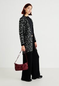 Cardigan s čiernym a šedým leopardím vzorom s dlhými rukávmi, kombinovaný so širokými čiernymi nohavicami a bordovou crossbody taškou s dvoma prednými vreckami.