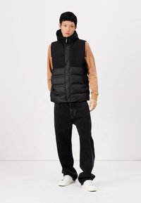 Svart puffervest med hette, front glidelås og trykknapper. Bært over en brun hettegenser, sammen med svarte jeans og hvite joggesko.
