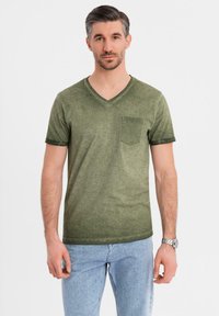 Olivově zelené tričko s výstřihem do V a přední kapsou, s krátkými rukávy a texturovanou látkou. V kombinaci se světle modrými džíny.