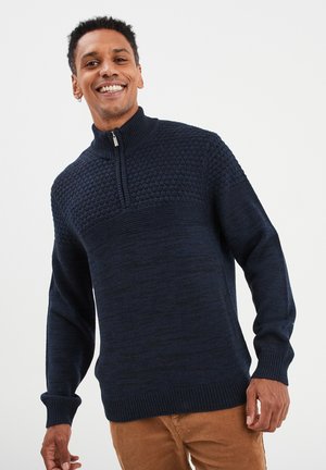 Jersey de punto azul marino con cuello alto y media cremallera, que presenta un patrón de punto texturizado en la parte superior del pecho y puños y dobladillo de canalé.