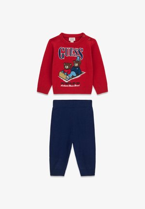 Raudona megzta džemperis su lokio grafika ir "GUESS" užrašu; derinamas su tamsiai mėlynomis megztomis kelnėmis su ažūrinėmis rankovėmis patogumui.