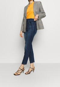 Blazer de cuadro gris, camiseta naranja con texto dorado, jeans azul de tiro alto y tacones negros con tiras. Ajuste relajado y estilo casual.