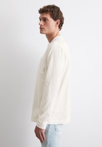 Langärmliges Baumwoll-T-Shirt in hellbeige mit einer Brusttasche, rundem Halsausschnitt und geradem Saum, kombiniert mit hellblauen Jeans.