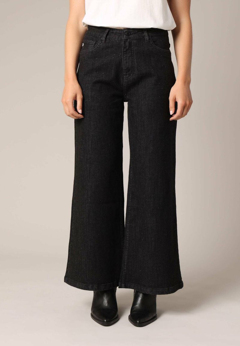Jean large noir en denim, avec une taille haute, fermeture à bouton sur le devant et un design classique à cinq poches. Associé à des bottines noires.