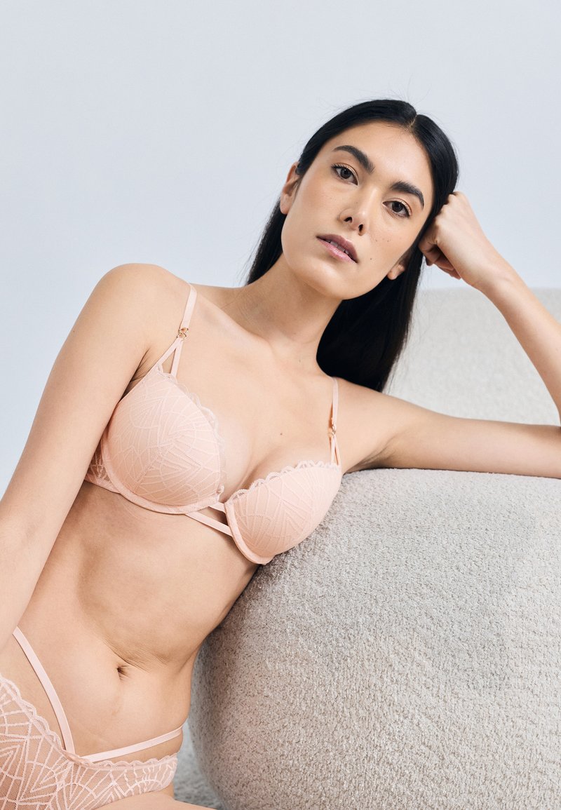 Őszibarackszínű csipkés melltartó formázott kosarakkal és hullámos szegélyekkel, vékony állítható pántokkal és geometriai mintával.