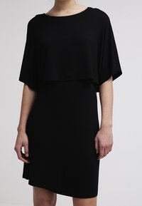Černé šaty s volnou, zavěšenou vrstvou a přiléhavou spodní částí. Měkká látka, kulatý výstřih a asymetrický detail spodního lemu.