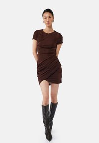 Robe marron à manches courtes avec des détails fronc�és, silhouette ajustée et design drapé. Assortie à des bottes noires longues. Texture lisse.
