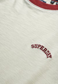 Camiseta de algodón blanca con un cuello acanalado color burdeos y el logo bordado "SUPERDRY" en el lado izquierdo del pecho, con un acabado de tejido texturizado.