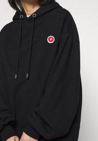 Sweat à capuche noir en tissu doux avec une poche kangourou à l'avant. Il comporte un patch circulaire rose avec un motif de beignet et des œillets argentés pour les cordons de serrage.