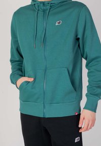 Teal zip-up hoodie med dragsko, framfickor, ribbad mudd och nederkant, med en liten svart logotyp på bröstet.