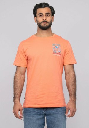 Homme barbu portant un T-shirt orange à imprimé tropical avec le texte "ST BARTH", associé à un jean bleu, debout devant un fond gris uni.