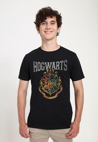 Černé bavlněné tričko s nápisem "HOGWARTS" v šedém textu a barevným erbem Bradavic uprostřed, s propracovanými detaily a zdobením.